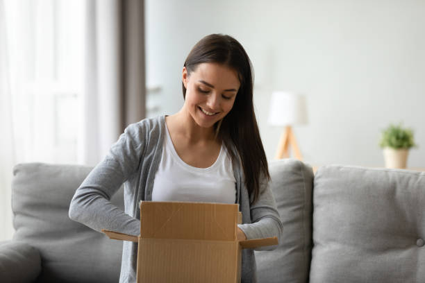 Satisfied woman unpacking box smiling enjoy fast delivery service Satisfied client young beautiful woman seated on couch in living room holding cardboard on lap feels pleased of delivery service unpacking box smiling enjoy moment of unbox purchased goods in web shop home delivery stock pictures, royalty-free photos & images