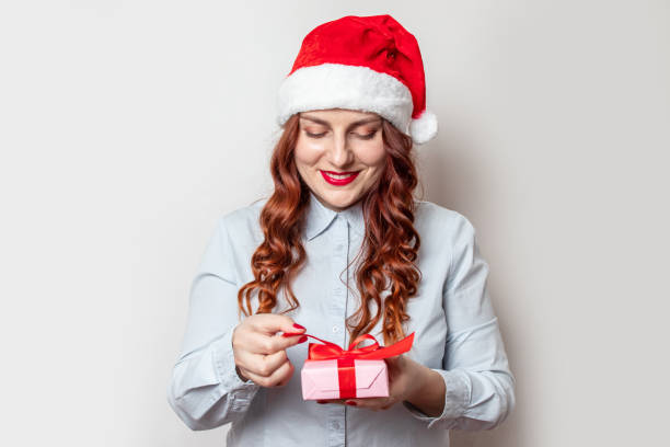 nöjd ung flicka med lockigt hår och en röd santa hatt på huvudet håller en presentask och ler mot en grå vägg - santa holding magazine bildbanksfoton och bilder