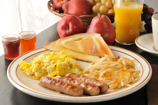 High angle shot of a country style scramble egg breakfast on a rustic wooden restaurant table. A skillet of hash brown potatoes is next to a blue plate with scrambled eggs, sausage links and toast.