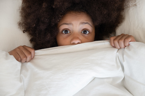 Scared cute little african kid girl looking at camera lying in bed cover with blanket, surprised small black child peeking from duvet wake from bad dream sleep feel fear afraid of nightmare, top view