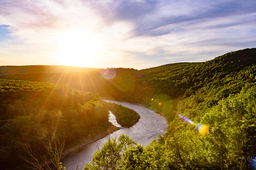 This is a photograph of the Delaware River winding through the Catskill Mountains and creating a natural state line between New York and Pennsylvania. The setting sun shines across the water and treetops in summer.