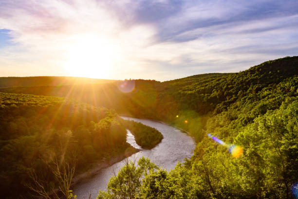 Ini adalah foto Sungai Delaware yang berkelok-kelok melalui Pegunungan Catskill dan menciptakan garis negara alami antara New York dan Pennsylvania. Matahari terbenam bersinar di atas air dan puncak pohon di musim panas.