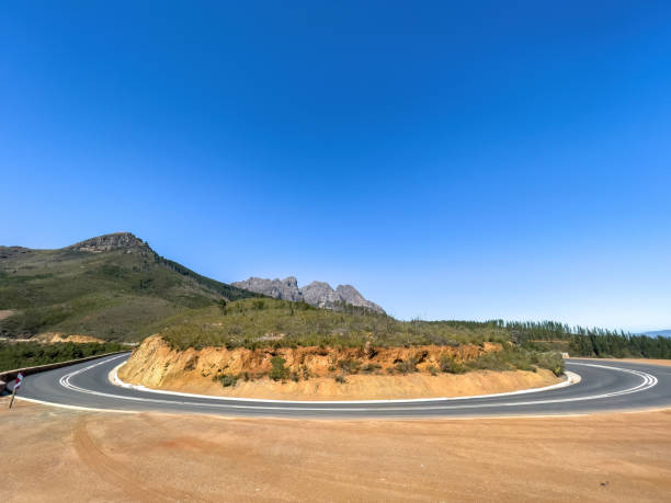 A scenic view of a road circling a rock outcrop, mountain peaks, and a vivid blue sky. stock photo