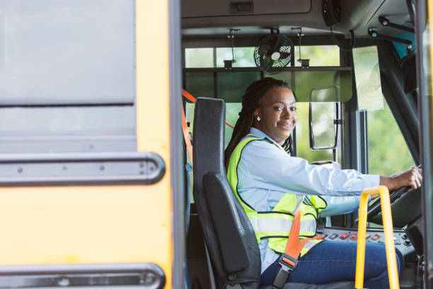 School bus driver looking through doorway A mature African-American woman in her 40s driving a school bus. She is sitting in the driver's seat wearing a reflective vest, looking toward the camera, through the open door. school bus driver stock pictures, royalty-free photos & images