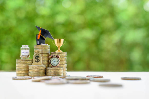 School funding, saving for higher education concept : Black graduation cap, campus diploma, US dollar bag, trophy cup, a clock on stacks of increasing coins. stock photo