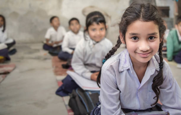 School girl in uniform of Indian Ethnicity sitting in their village classroom, looking at camera smiling. Girl sitting on the floor of her government primary school in uniform along with some of her class mates sitting behind her.  Selective focus, shallow depth of field. india stock pictures, royalty-free photos & images
