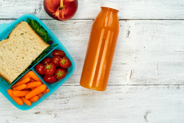 School lunch - plastic box with sandwich, tomatoes, carrots in sections with orange juice in bottle on white wooden background stock photo
