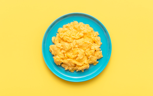 Scrambled eggs plate minimalist on a yellow table, view from above. Delicious homemade scrambled eggs in a blue plate isolated on a yellow background.