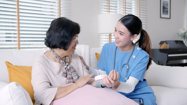 Scrub nurse doctor using digital tonometer check blood pressure for old asia female at home in elderly care cardiovascular medical visit, cholesterol problem and hospice healthcare caregiver concept. stock photo