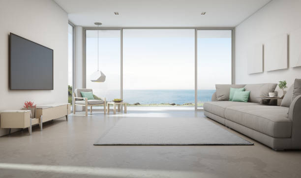 Sea view living room of luxury summer beach house with large glass door and wooden terrace. TV on white wall against big gray sofa in vacation home or holiday villa. 3d rendering of hotel interior. coastal feature stock pictures, royalty-free photos & images