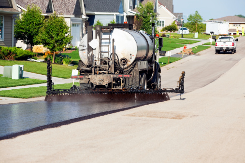 A spray truck is applying a black asphalt (tar) seal coat to a street. After this crushed rock will be applied to chipseal the road which is less costly resurfacing alternative. The bottom right, opposite side, of the street has already received a coating of rock.