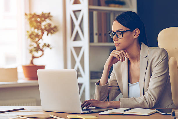 Searching for new solution. Pensive young beautiful businesswoman in glasses working on laptop and keeping hand on chin while sitting at her working place hand on chin stock pictures, royalty-free photos & images
