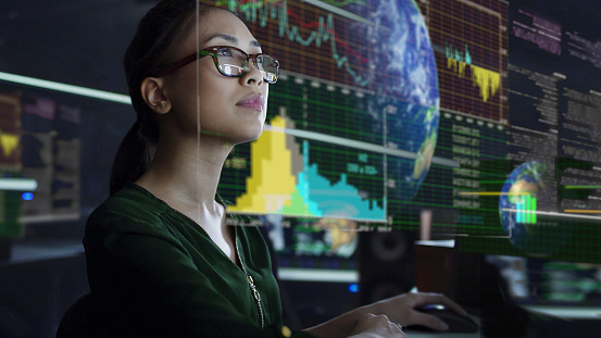 Stock photo of a young Asian woman looking at see through global & environmental data whilst seated in a dark office. The data is projected on a see through (see-thru) display.