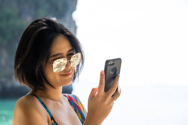 Selective focus at the face of young Asian women, look and smile at her smartphone while travel on summer vacation beach. Mobile wireless communication for social media or real time online call. Selective focus at the face of young Asian women, look and smile at her smartphone while travel on summer vacation beach. Mobile wireless communication for social media or real time online call. real time footage stock pictures, royalty-free photos & images