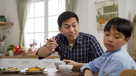 selective focus frustrated Asian father holding a chopsticks with vegetable and asking his boy son to take a bite in dining room at home. the boy turns head away and refuses to eat