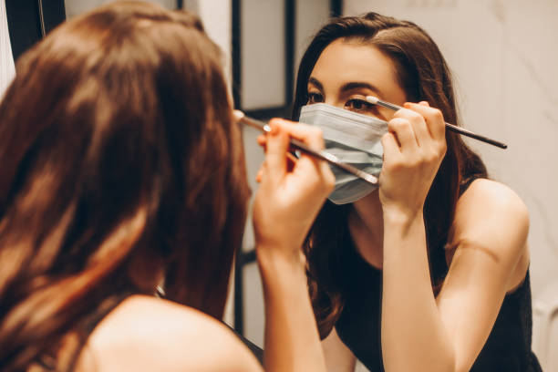 selective focus of young woman in medical mask and black dress applying eye shadow in bathroom - make up imagens e fotografias de stock