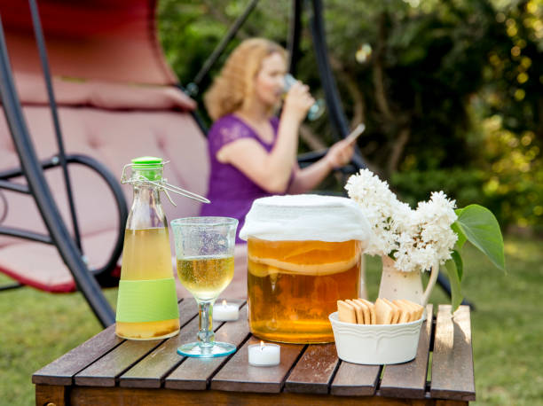 Selective focus on jar with kombucha drink and tea mushroom in it and served in goblet glass outdoors in sunny summer evening, blurred tea drinking woman on the background. Natural probiotics concept.  kombucha stock pictures, royalty-free photos & images
