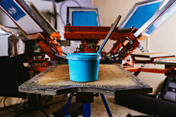 selective focus photo of can of blue paint on wooden shelve on the print screening apparatus. serigraphy production. printing images on t-shirts by silkscreen method in a design studio stock photo