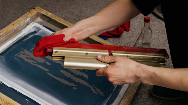selective focus photo of hands with squeegee. serigraphy production. printing images on t-shirts by silkscreen method in a design studio stock photo