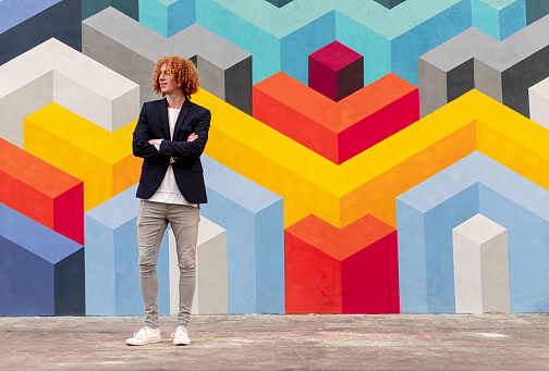 Full body of cheerful young stylish male with re curly hair in trendy outfit standing on street with crossed arms and looking away, against colorful geometric graffiti wall