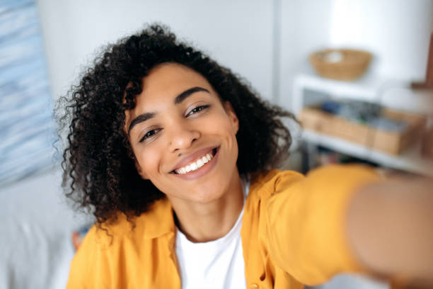 Selfie photo on smartphone. Joyful carefree curly african american girl, student, freelancer, makes a selfie on the phone, fooling around, having fun, looks at the phone's webcam, smile happily stock photo