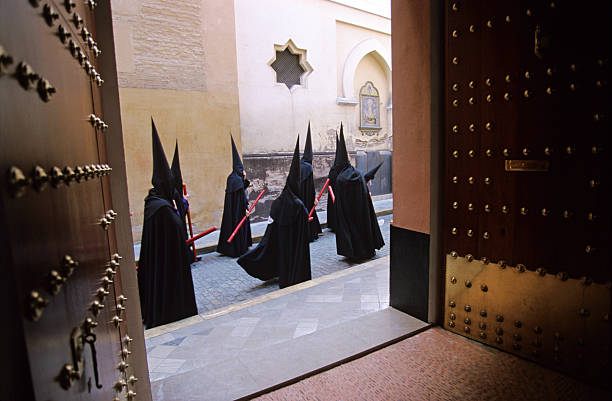 Semana Santa in Seville "Holy week in Seville (Spain) is marked by citizens dressed in penitential robes with tall hoods to disguise their identity -  parading through the streetsHere a few of the hundreds of pilgrims pass a church in procession  (a Christian emblem is visible on the wall) and are framed in a gatewayHoly Week in Seville (Semana Santa de Sevilla) is one of the most important traditional events of the city. It is celebrated in the week leading up to Easter (Holy Week among christians), and is one of the better known religious events within Spain." holy week stock pictures, royalty-free photos & images