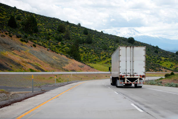 Semi truck with semi trailer move down on winding highway with green hills A big rig semi truck with a dry van trailer for long haul freight turn on winding dividing highway with green trees on roadside hills semi truck back stock pictures, royalty-free photos & images