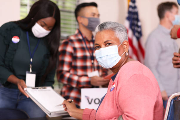 Senior adult, African descent woman votes in USA election wearing mask. African descent, senior adult woman votes in the USA election wearing a protective mask to protect herself against COVID-19 or other infectious diseases.  She is in a wheelchair. polling place stock pictures, royalty-free photos & images