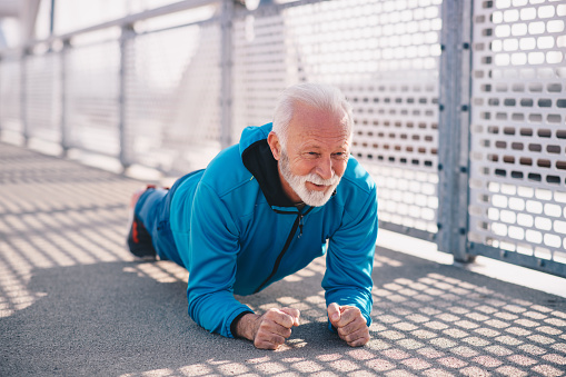 A senior adult planking outdoors. He is wearing blue sports clothing. It's a sunny day.