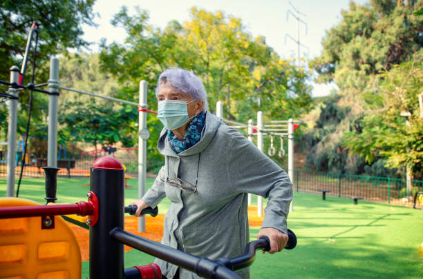 A senior adult woman in a mask starts exercising in an outdoor fitness field near her residence. stock photo