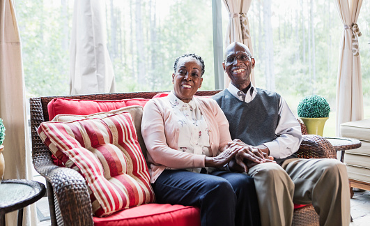 A senior African-American couple sitting together at home on a couch. They are holding hands, smiling at the camera. They are both in their 70s.