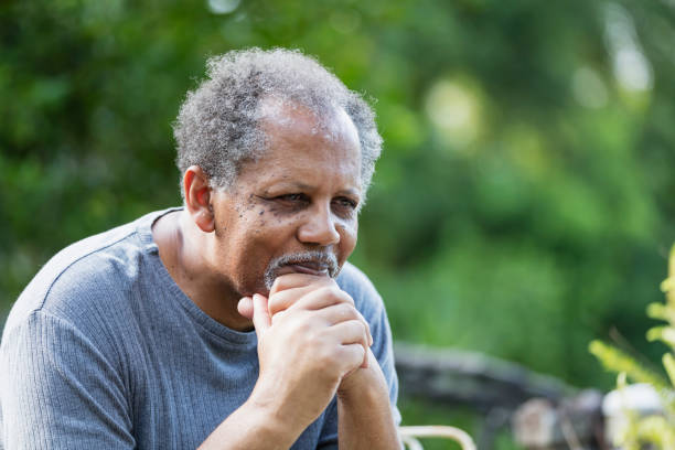 Senior African-American man outdoors, thinking A senior man sitting outdoors in his back yard with a sad expression on his face, leaning his chin on his clasped hands. He is an African-American man in his 60s. sad old black man stock pictures, royalty-free photos & images