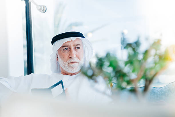 Senior Arabian Man with cellphone Portrait of senior Arabic businessman wearing traditional emirati clothes (dishdasha) sitting, looking at camera and holding smart phone at modern cafe restaurant in shopping mall. old arab man stock pictures, royalty-free photos & images