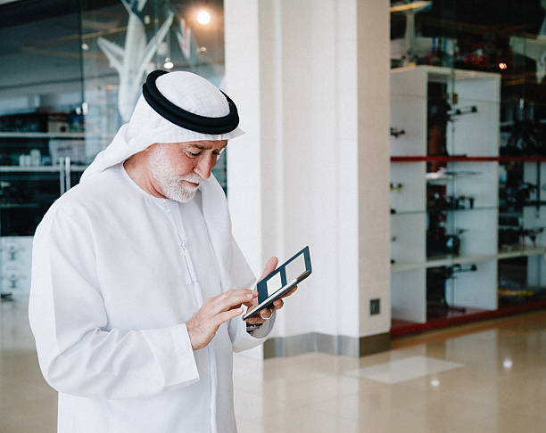 Senior Arabian Man with cellphone Portrait of senior Arabic businessman wearing traditional emirati clothes (dishdasha) holding smart phone and texting at modern  shopping mall. old arab man stock pictures, royalty-free photos & images