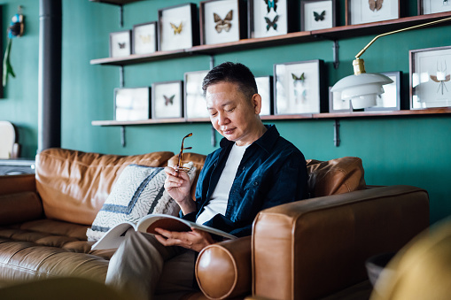 Senior Asian man sitting on sofa in the living room, relaxing and reading a book at home