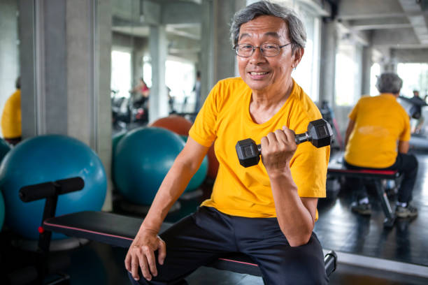 Senior asian sport man lifting dumbbells in fitness gym . elder male exercising ,  working out , training weights, healthy ,Retirement , older, looking camera stock photo