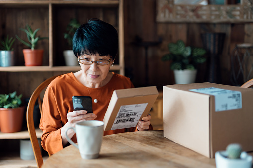 Senior Asian woman relaxing at home, shopping online with smartphone, receiving delivered parcels by home delivery service. Online shopping, online banking. Enjoyable customer shopping experience