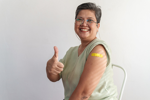 Portrait of senior Asian woman with eyeglasses sitting against white background, giving thumbs up and showing her arm with yellow bandage after got vaccinated, smiling and looking at camera.