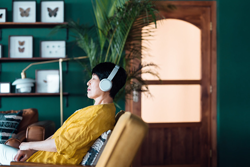 Senior Asian woman with eyes closed, enjoying music over headphones while relaxing on the armchair at home