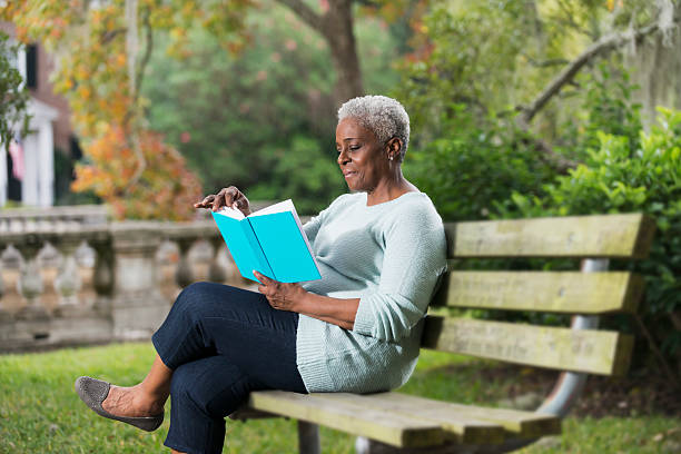 Senior black woman reading a book A senior African American woman in the park, sitting on a bench reading a book. park bench stock pictures, royalty-free photos & images