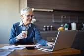 Female senior business woman using laptop at home office. Mature gray hair manager using computer while drinking coffee