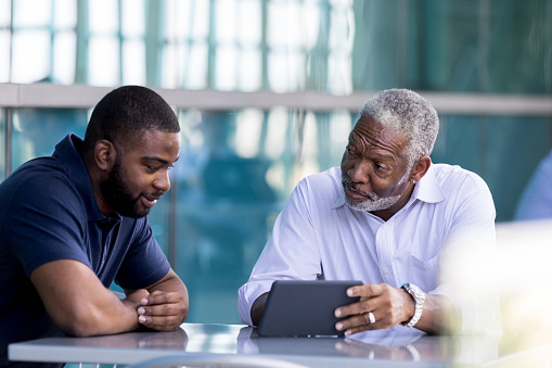 A senior African American businessman shows a male colleague something on a digital tablet while sitting outside on an office patio.