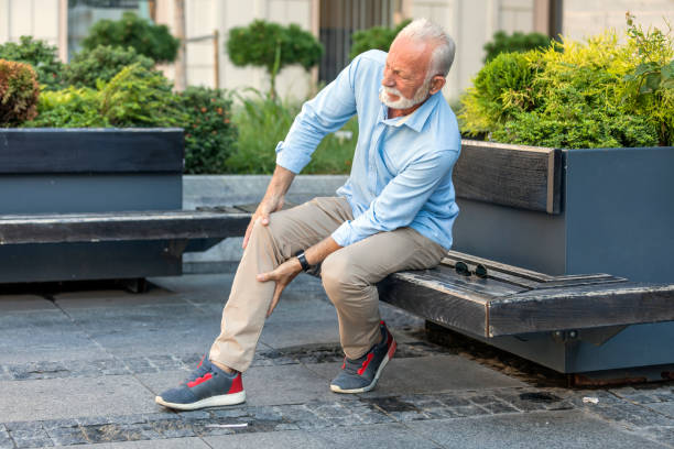 Senior Businessman with Knee Problems in the City Streets Mature Man with Grey Hair and Beard has Problems with his Knee in the City. Older Man is Sitting on the Wooden Bench in Public Park and Holding his Knee due to Physical Injury. knee stock pictures, royalty-free photos & images