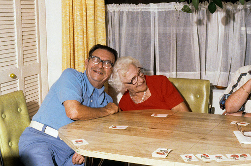A horizontal image of cheerful senior citizens who are playing cards at the kitchen table.  They are both laughing and leaning in together. It is a genuine happy and cheerful image.