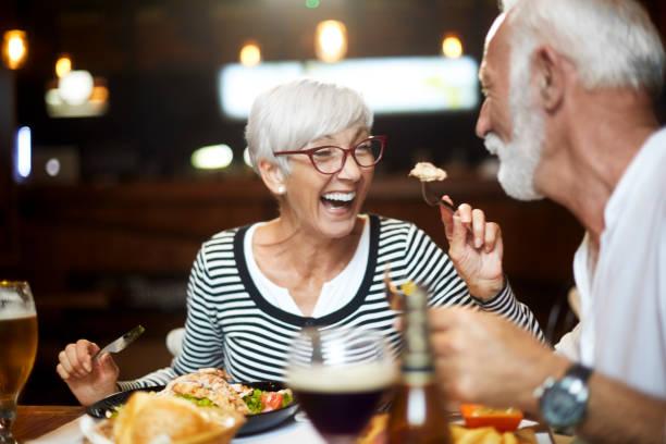 senior paar voeden elkaar en met een goede tijd tijdens een maaltijd in een restaurant - avondmaaltijd stockfoto's en -beelden