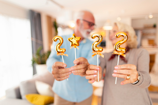 Cheerful senior couple holding golden balloons shaped as numbers 2022, representing the upcoming New Year