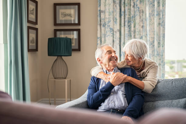 Senior couple hugging and having fun at home Happy senior woman embracing her husband at home while laughing together. Smiling wife hugging old man sitting on couch from behind. Joyful retired couple having fun at home while looking at each other with copy space. Love and unity concept. 70 79 years stock pictures, royalty-free photos & images