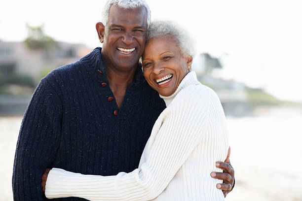 Senior couple sharing a romantic hug on the beach Romantic Senior Couple Hugging On Beach Looking At Camera Laughing old black couple in love stock pictures, royalty-free photos & images
