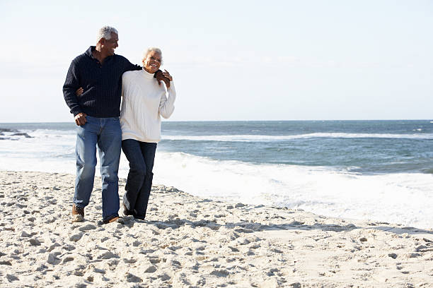 Senior Couple Walking Along Beach Together Senior Couple Walking Along Beach Together With Arms Round Each Other Smiling old black couple in love stock pictures, royalty-free photos & images