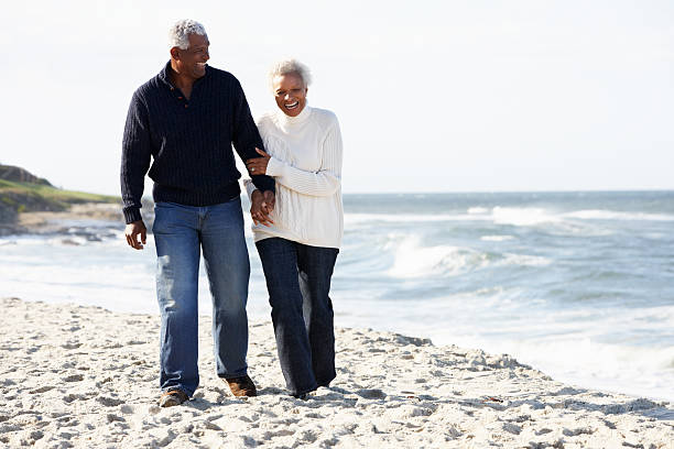 Senior couple walking along the beach while holding hands Senior Couple Walking Along Beach Together Holding Hands Laughing old black couple in love stock pictures, royalty-free photos & images